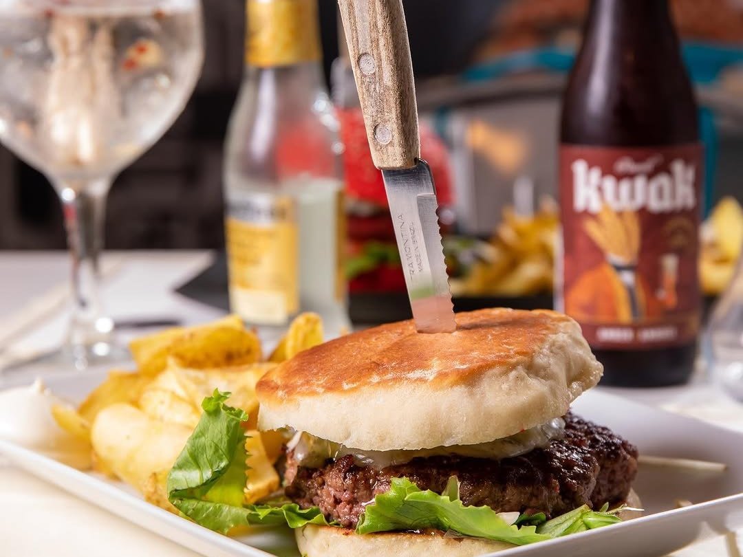 Hambúrguer de carne no bolo do caco, com alface e batatas fritas, ao lado de um copo e de uma garrafa de cerveja
