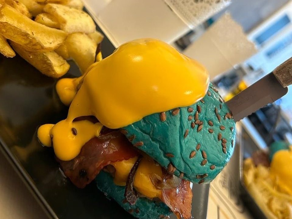 Hambúrguer em pão azul com sementes, coberto de queijo derretido, ao lado de batatas fritas