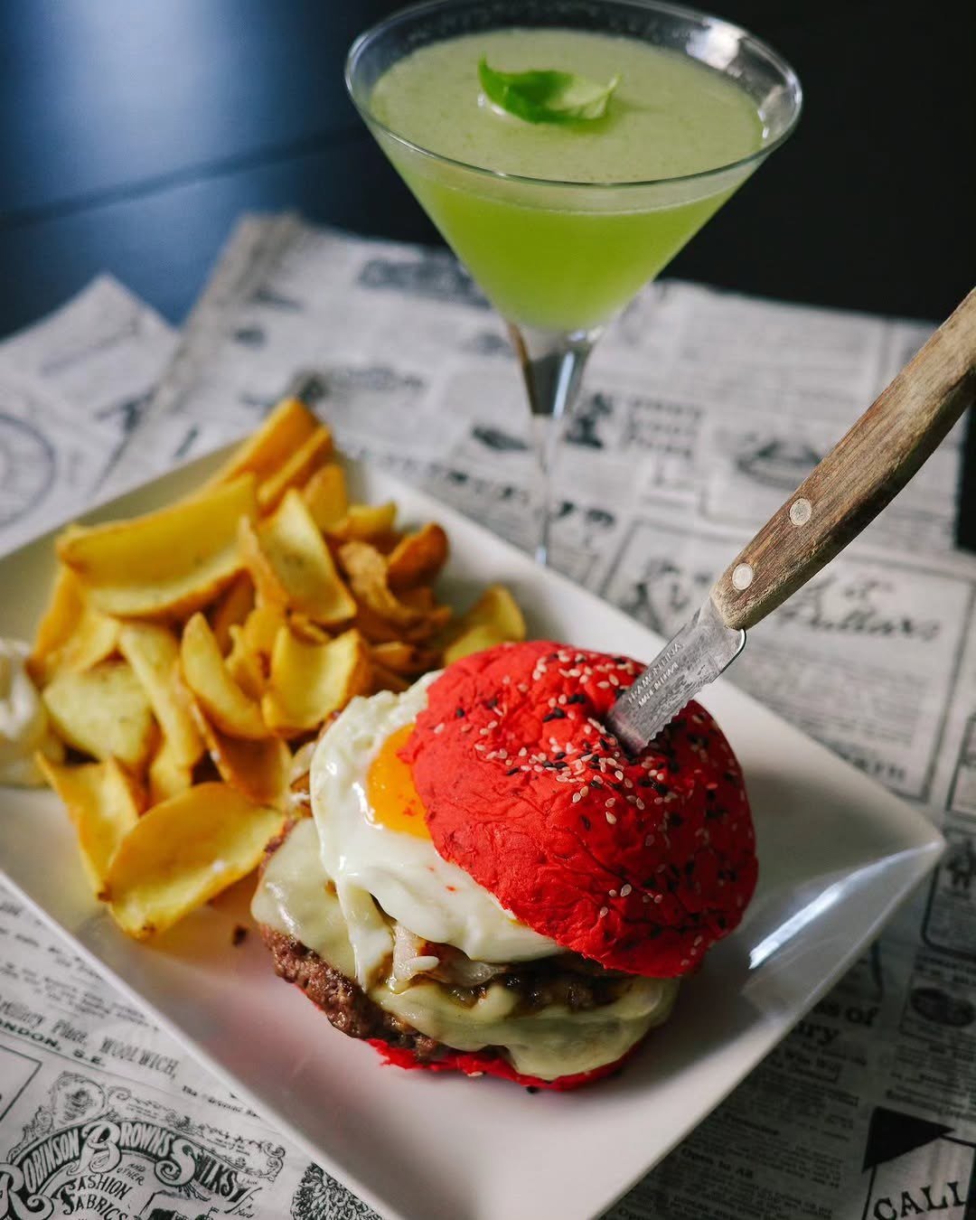 Hambúrguer em pão vermelho com ovo estrelado e batatas fritas, servido sobre papel de jornal, com um cocktail verde ao fundo