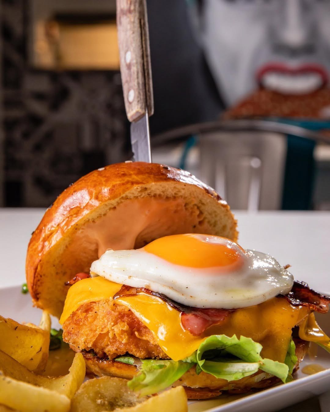Hambúrguer de frango panado em pão tostado, com ovo estrelado, queijo cheddar, bacon e alface, ao lado de batatas fritas